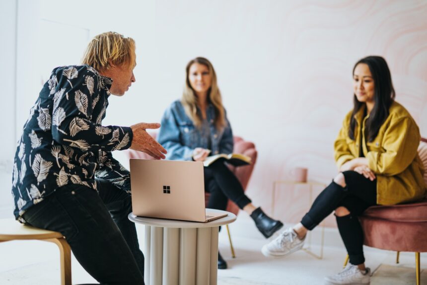 man in black and white floral long sleeve shirt sitting and showing something on a microsoft laptop to 2 women